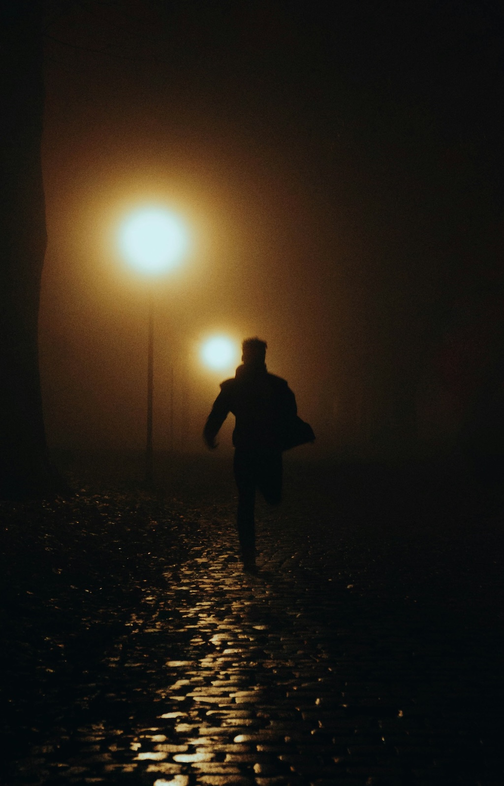 A person, KarmaMan, running on a wet cobblestone path at night, illuminated by streetlights through fog.